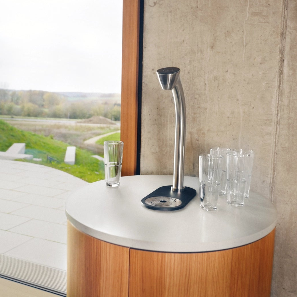 A modern stainless steel BRITA EXTRA C-Tap water dispenser with a curved neck and black base, set on a round wooden and white countertop next to clear drinking glasses, with a concrete wall and outdoor patio visible in the background.