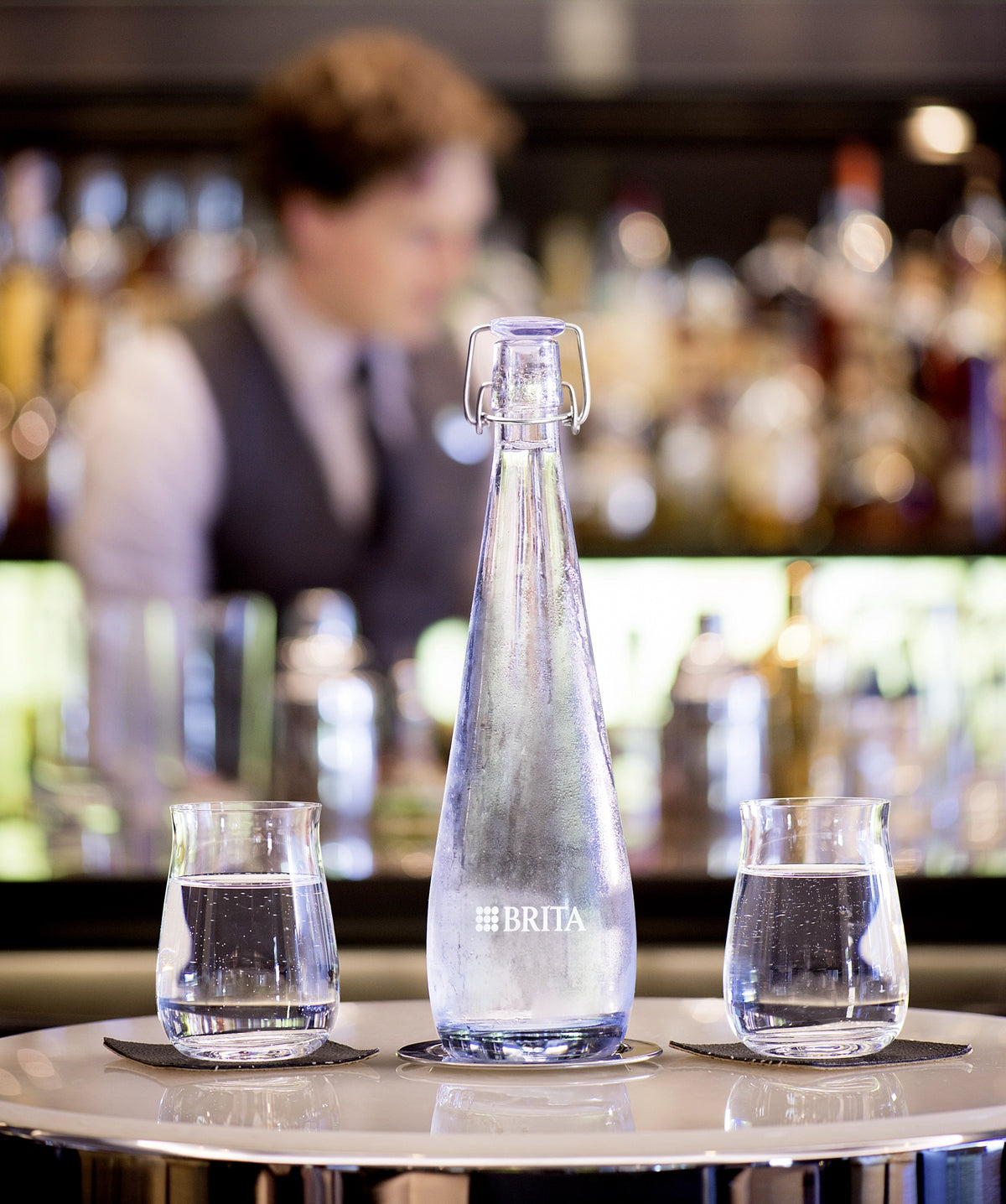 Een BRITA Swing-fles met twee glazen water op een bar, met op de achtergrond een wazige barman.