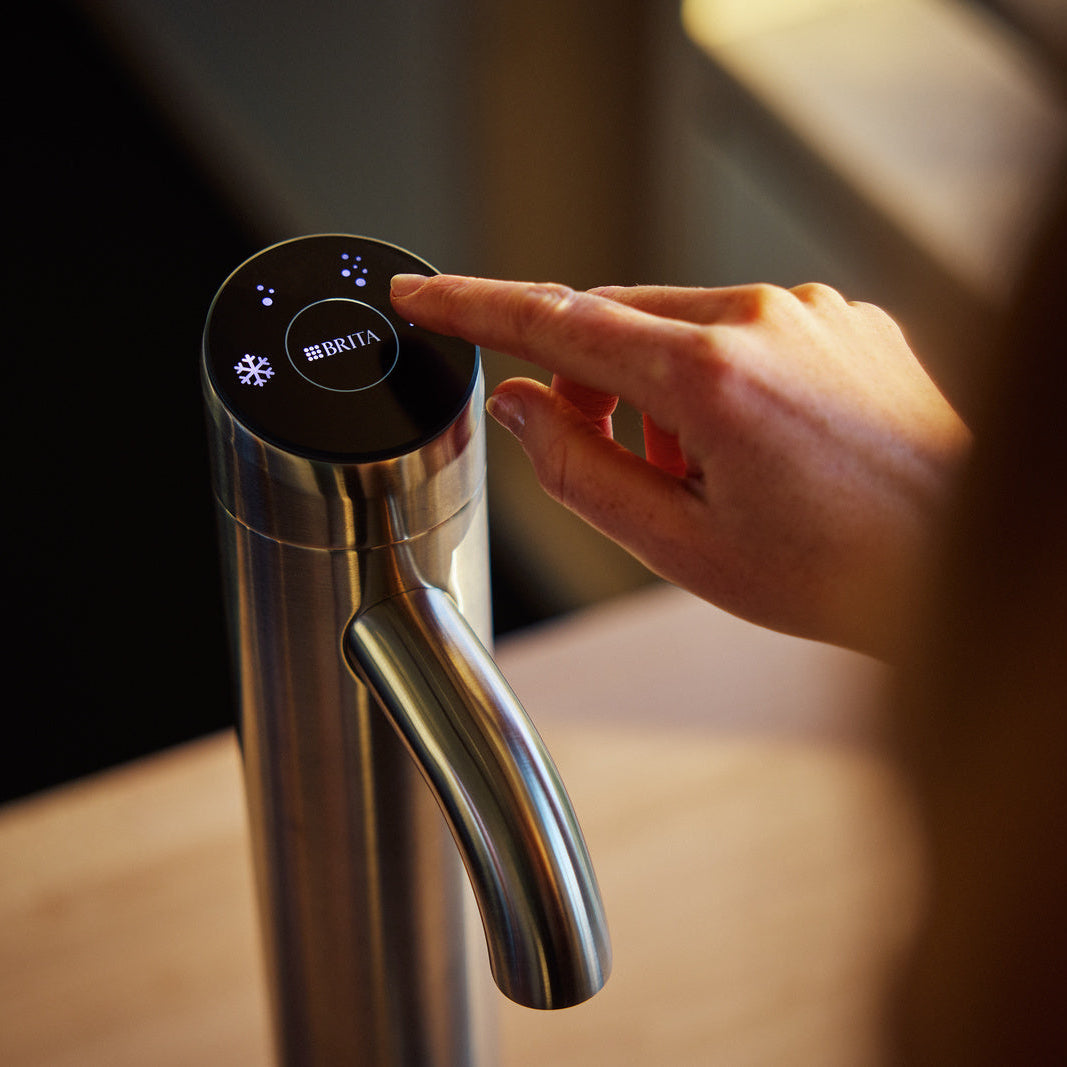 A close-up of a stainless steel BRITA Extra C-Tap water dispenser with a black touch control panel, being operated by a person's finger in a warm indoor setting.